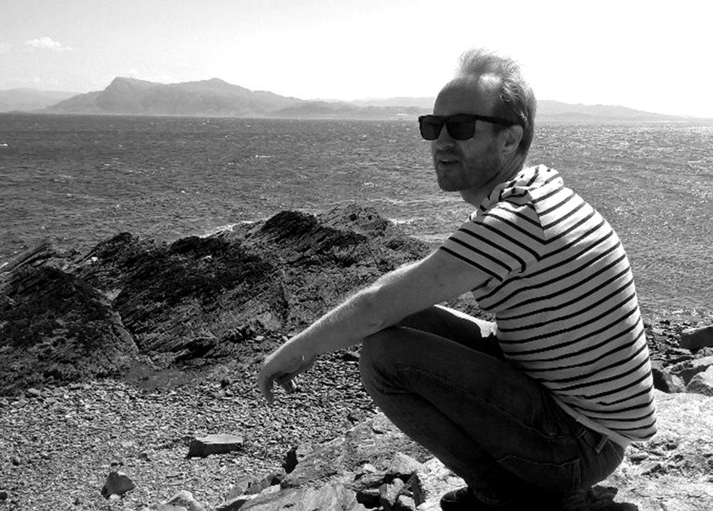 Neil crouching, happily, on a wall on the side of a Loch in Scotland. The sun is shining, the mountains are misty in the background. All is well. Silence all around.