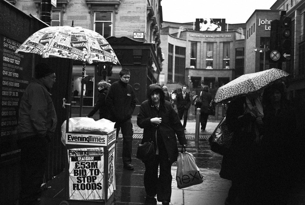 A crowd scene on Glasgow's Buchanan Street. In sudden rain, a lady with a hood up walks past a newspaper seller who's board reads "53M Pounds Bid to Stop Floods". In the background, there is a street sign reading "Bath Street".