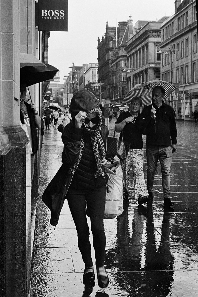 In a torrential downpour of rain, a lady, wearing sunglasses, holding a shopping bag, and pulling her hood down to shield her from the rain, rushes towards the camera